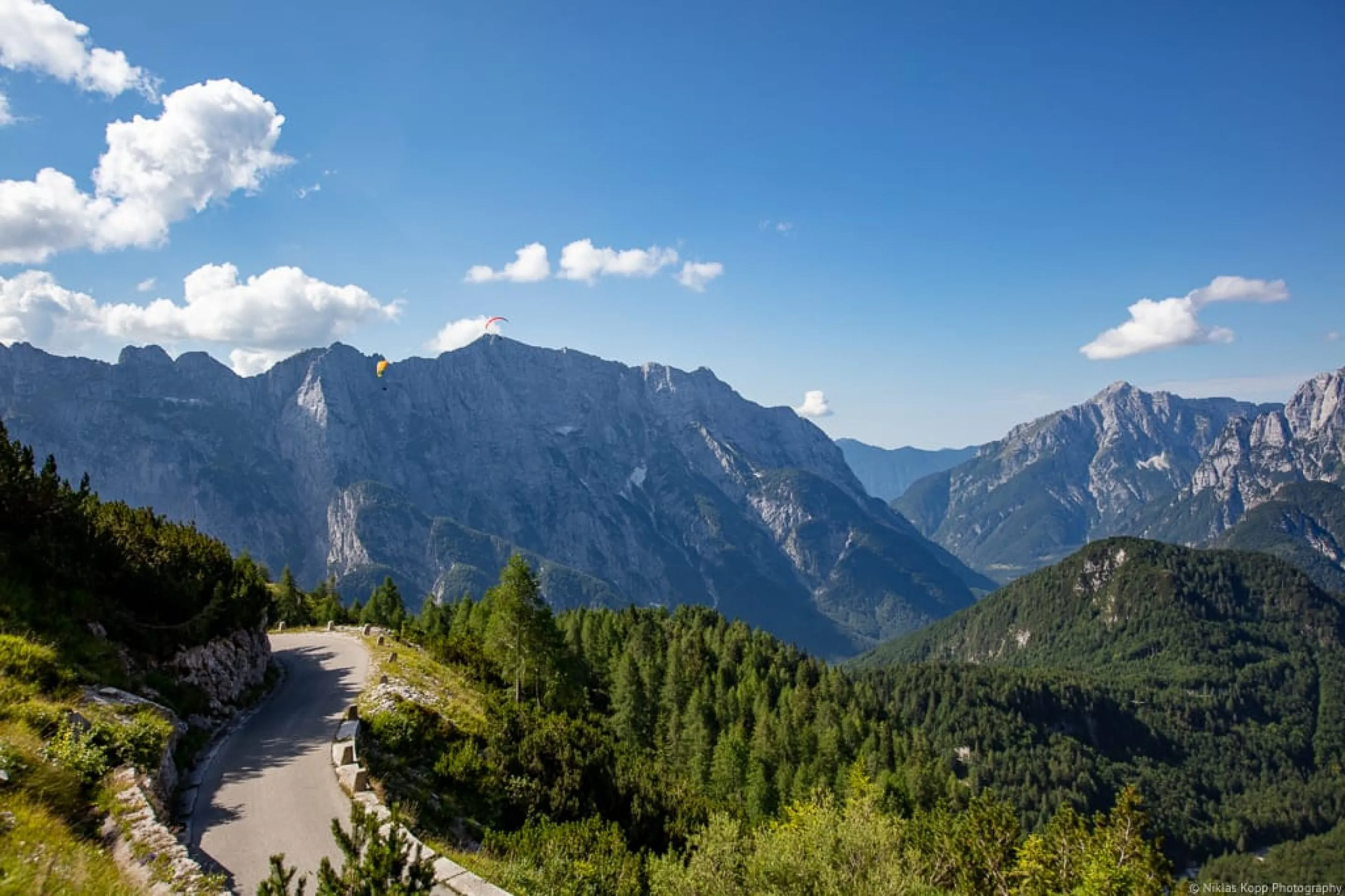 A narrow mountain road with paragliders in the sky